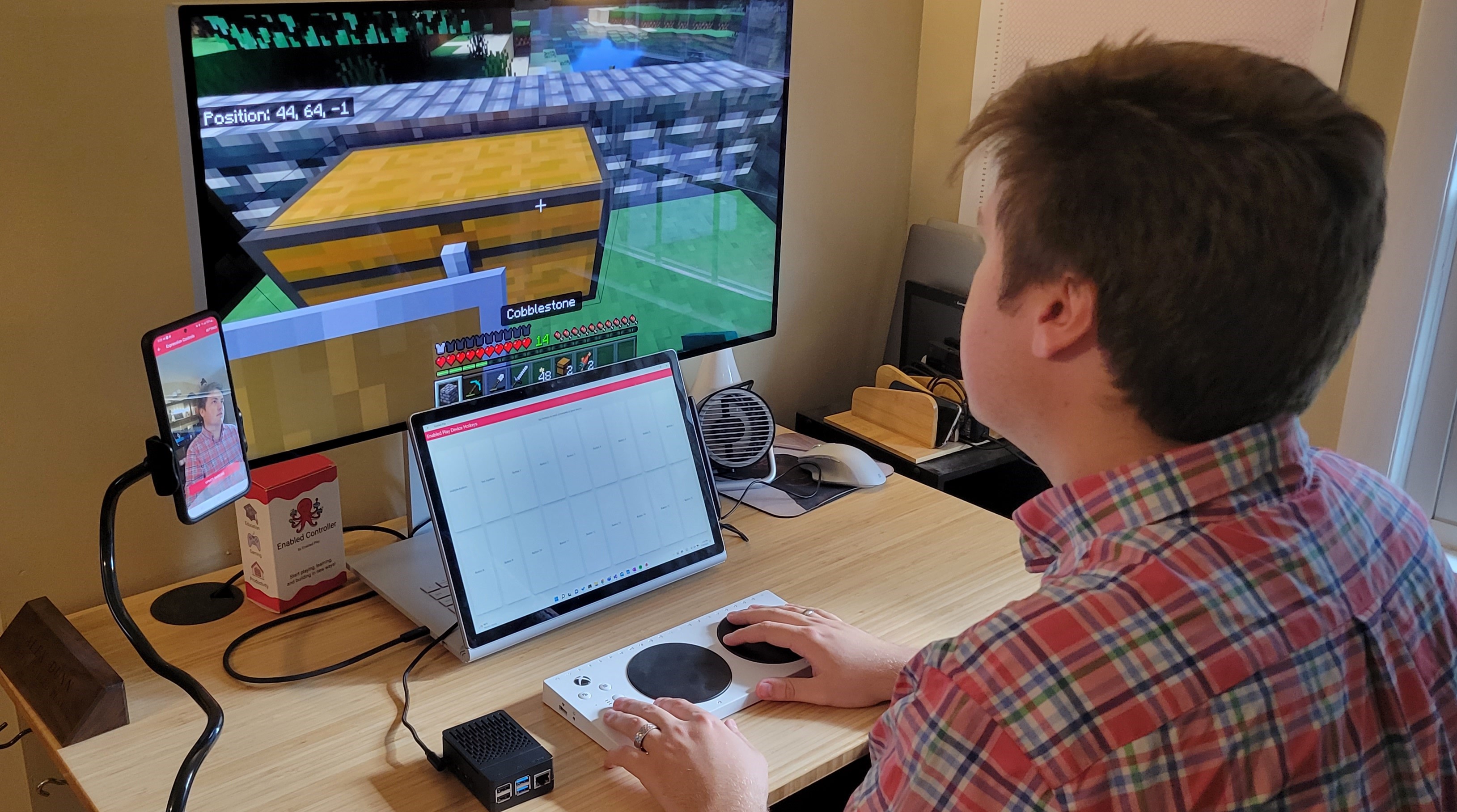 A young person sitting at a desktop computer playing the game, Minecraft, using an accessible controller to control the movement of the character.